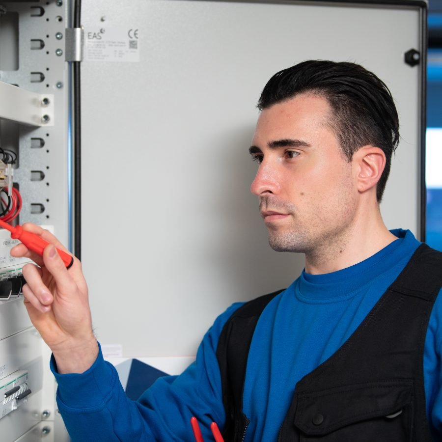 En elektriker i gjenkjennelig Norgeseliten-blå genser monterer strømbryteren i sikringsskapet.